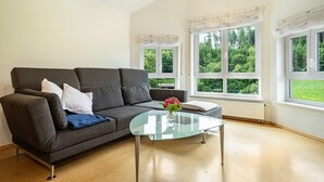 Ein modernes Wohnzimmer mit einem grauen Sofa, einem Glastisch und großen Fenstern mit Vorhängen.