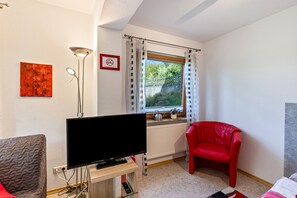 A room with a television on a stand, a red chair, a red couch, a window with curtains, a radiator, and a wall-mounted artwork.