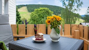 Une part de gâteau sur une assiette, posée sur une table, avec un vase de fleurs et une vue sur un paysage verdoyant.