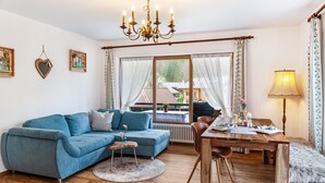Un salón con un sofá azul, una mesa de comedor de madera, una araña y vistas a una casa a través de amplios ventanales.
