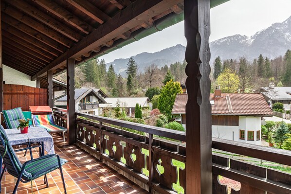 Un balcón techado con barandilla de madera, una mesa con sillas y vistas a un paisaje monta ñoso.