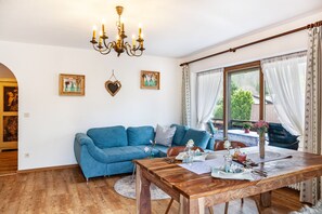 Una sala de estar con un sofá azul, una mesa de comedor de madera, un candelabro y ventanas grandes con cortinas.