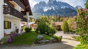 A mountain lodge with a stone pathway, wooden balconies, and a view of snow-capped mountains. - Studio Apartment 'Aschaubichl - Anemone' with Mountain View, Balcony and Wi-Fi (Grainau)