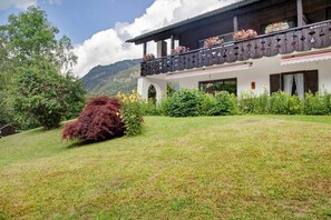 A traditional chalet with a balcony adorned with flowers, surrounded by lush greenery and mountains in the background.