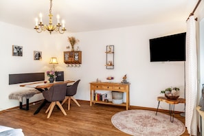 A modern living room with a wooden floor, a dining table with chairs, a TV mounted on the wall, and a chandelier.