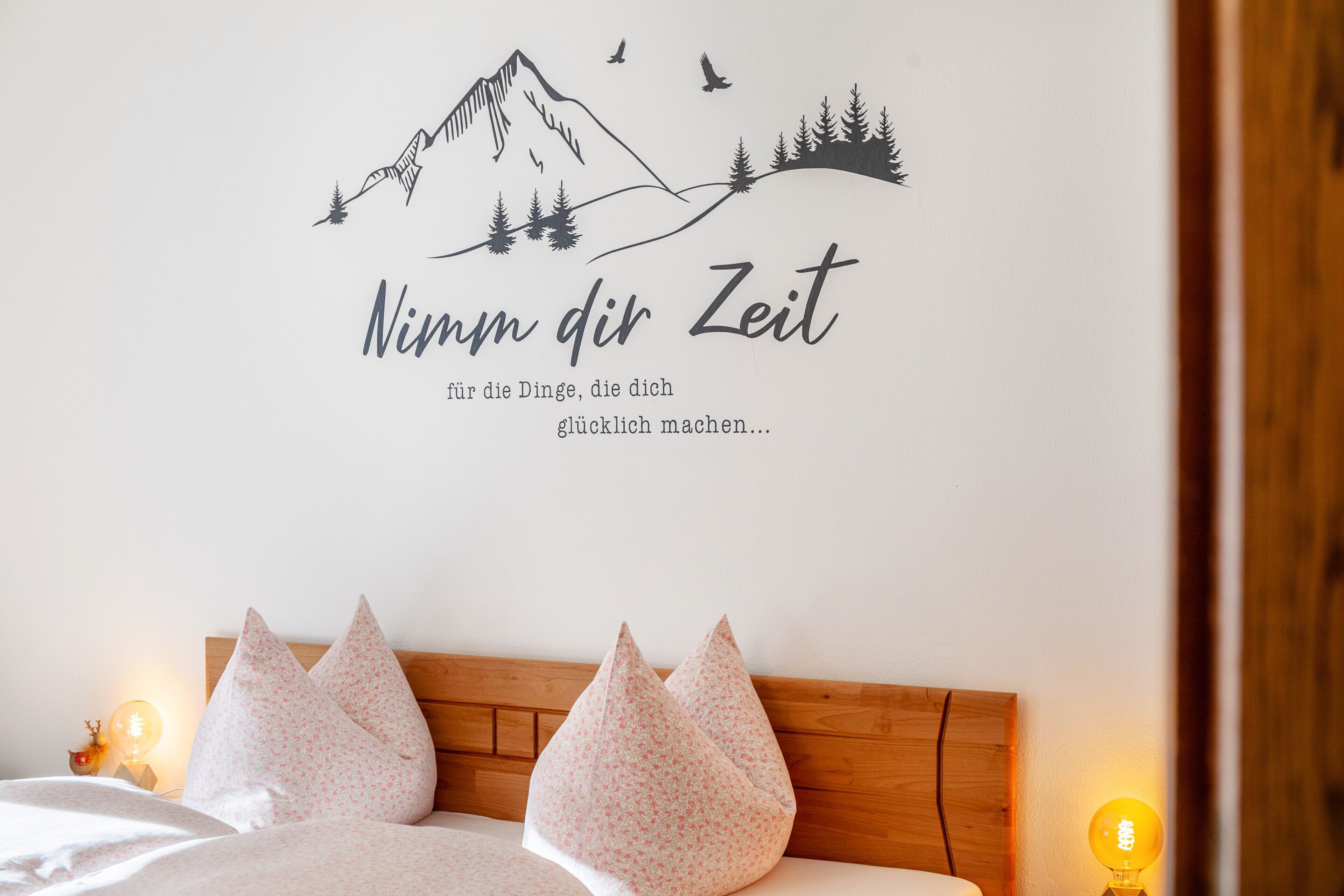 A bedroom with a mountain-themed wall art, a wooden headboard, and two bedside lamps.