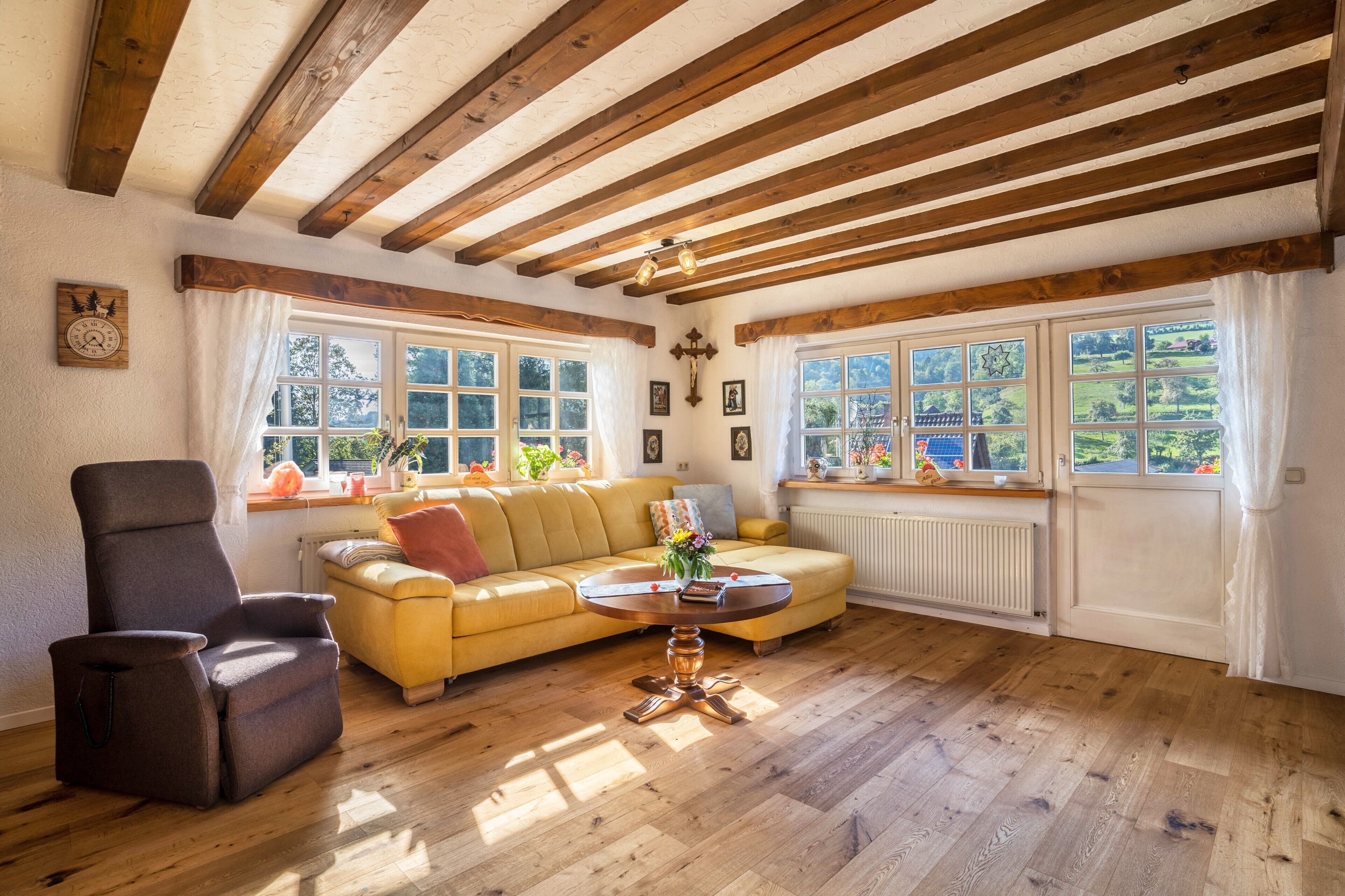 A room with wooden beams, a sofa, a chair, a table with flowers, and large windows with curtains.