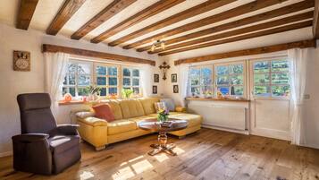 A room with wooden beams, a sofa, a chair, a table with flowers, and large windows with curtains.