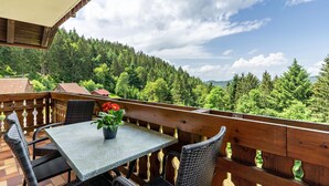 A balcony with a table and chairs, a potted plant, and a view of a forested hillside. - Holiday Apartment "Fuchsmichelhof 2" (Seebach)