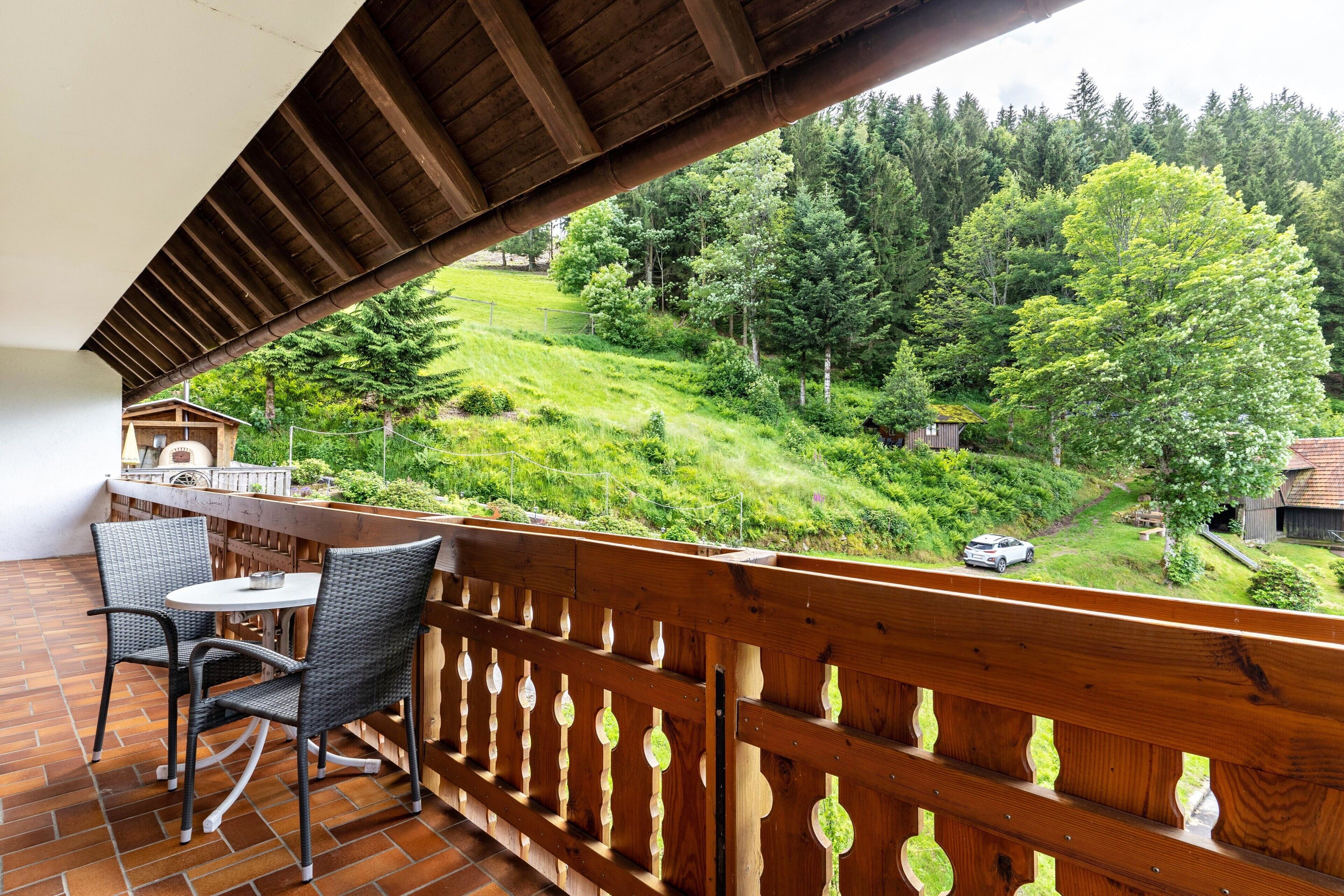 Un balcon avec des fauteuils en osier et une table, donnant sur une colline verdoyante parsemée d’arbres et où l’on aperçoit une voiture.