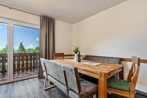 Een eetgedeelte met een houten tafel, stoelen met kussens, een vaas met bloemen en uitzicht door een glazen deur.