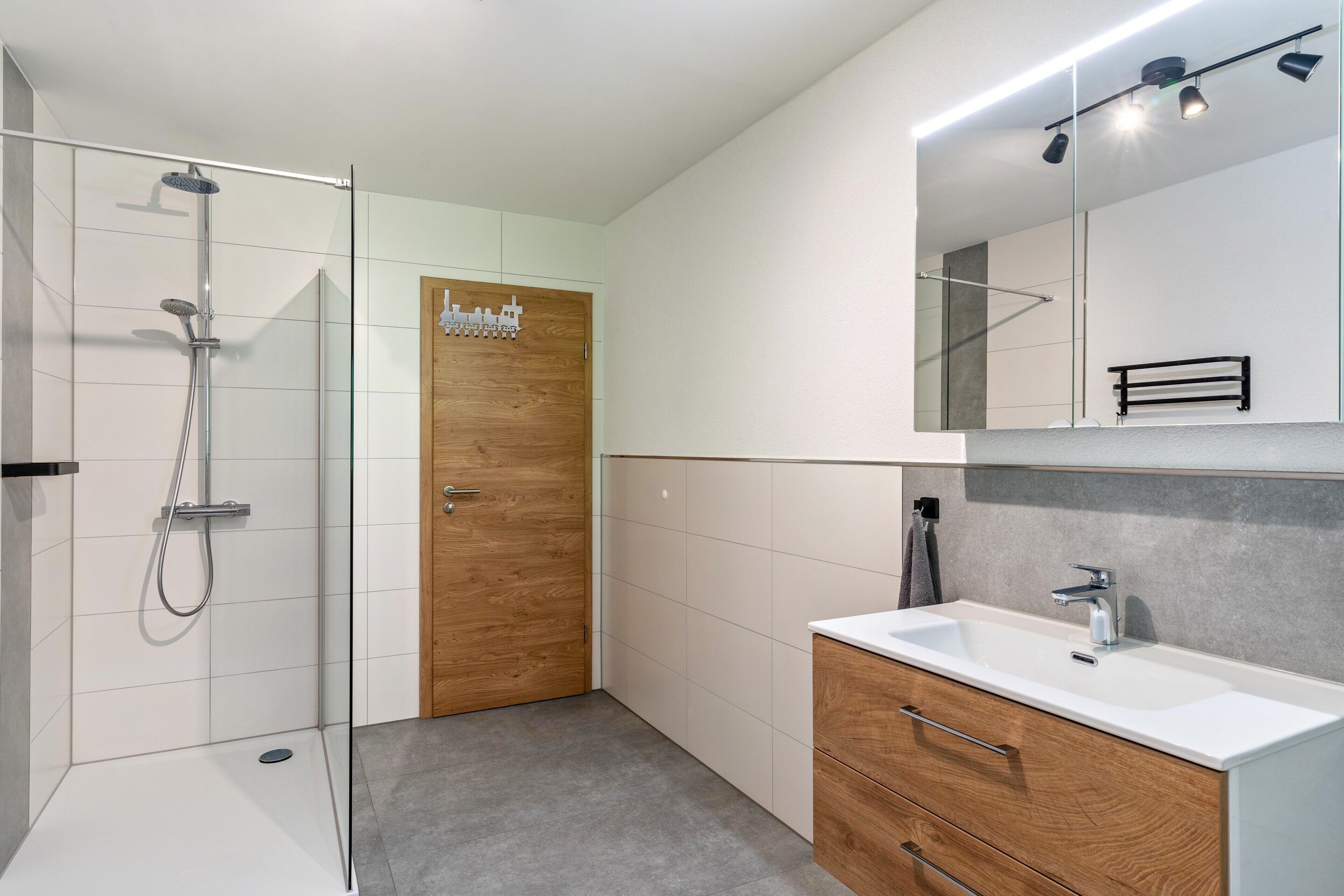 Une salle de bain moderne avec une douche à parois de verre, une porte en bois, un lavabo blanc avec un miroir au-dessus et un porte-serviettes.