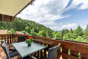 A balcony with a table and chairs, a potted plant, and a view of a forested hillside.