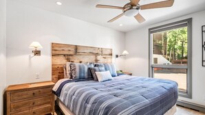 A bedroom with a large bed, wooden headboard, two bedside lamps, a ceiling fan, and a window with a view of trees. - Perfect Location Between Vail & Loinshead (Vail)