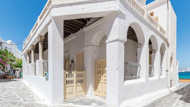 Un edificio blanco con un acceso cubierto, calle empedrada de piedra y vista al mar.