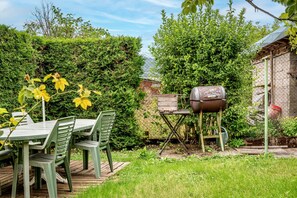 Une terrasse avec une table et des chaises, un barbecue et un jardin agrémenté de haies et d’arbres.