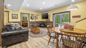 A cozy living room with a fireplace, leather sofa, wooden dining table, and a TV mounted on the wall. - Accessible Lake and Farm Retreat w/Fresh Eggs, Nature Trails, and Gameroom (Thornfield)