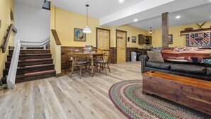 A spacious living area with a wooden staircase, a dining table with chairs, a sofa, and a patterned rug. - Accessible Lake and Farm Retreat w/Fresh Eggs, Nature Trails, and Gameroom (Thornfield)