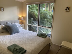 A bedroom with a bed, pillows, a nightstand with a lamp, a window with a view of trees, and a chair. - Sassafras Treehouse in The Dandenong Ranges, Victoria (Sassafras)