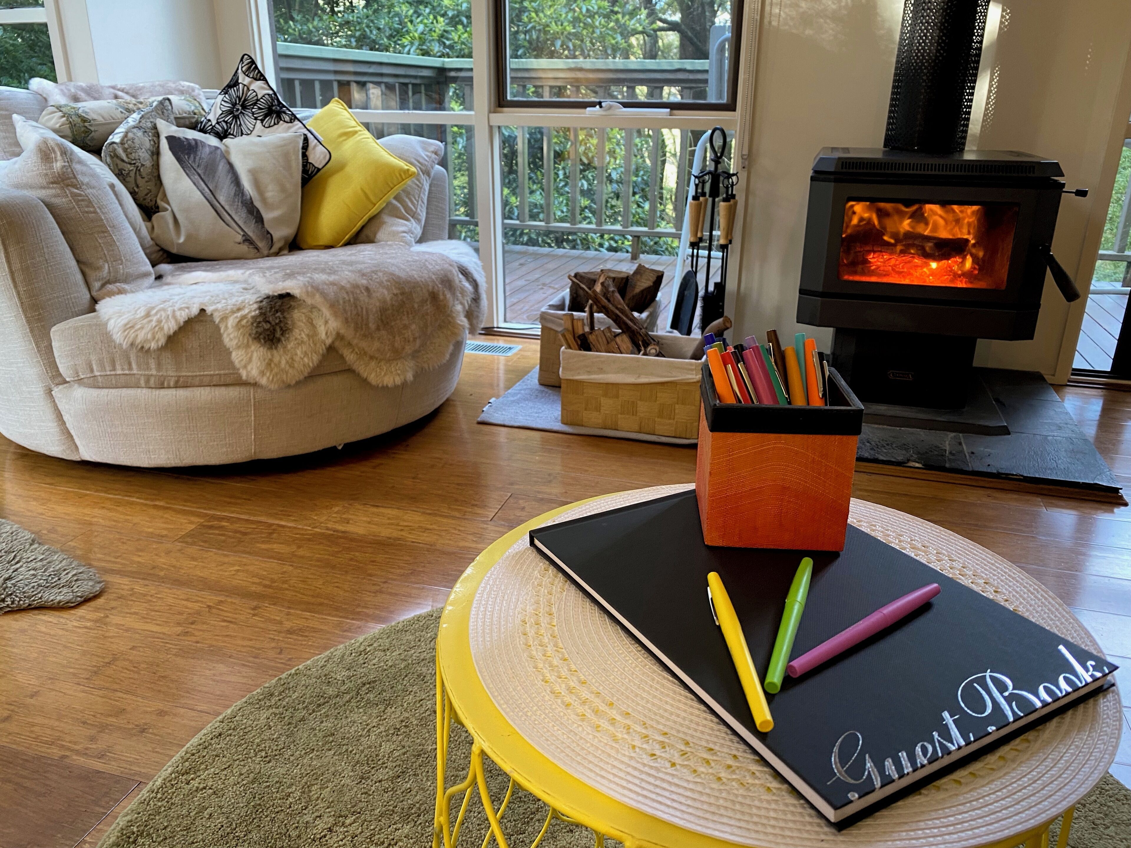 A cozy living room with a round yellow table, a black notebook labeled "Guest Book," and a colorful pen holder.