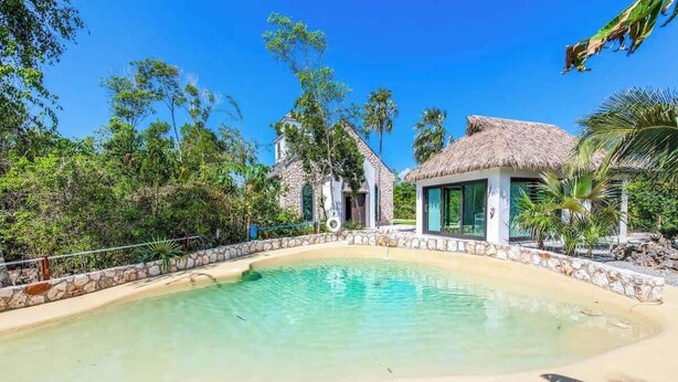 A pool surrounded by stone walls and tropical vegetation, with a thatched-roof building nearby.