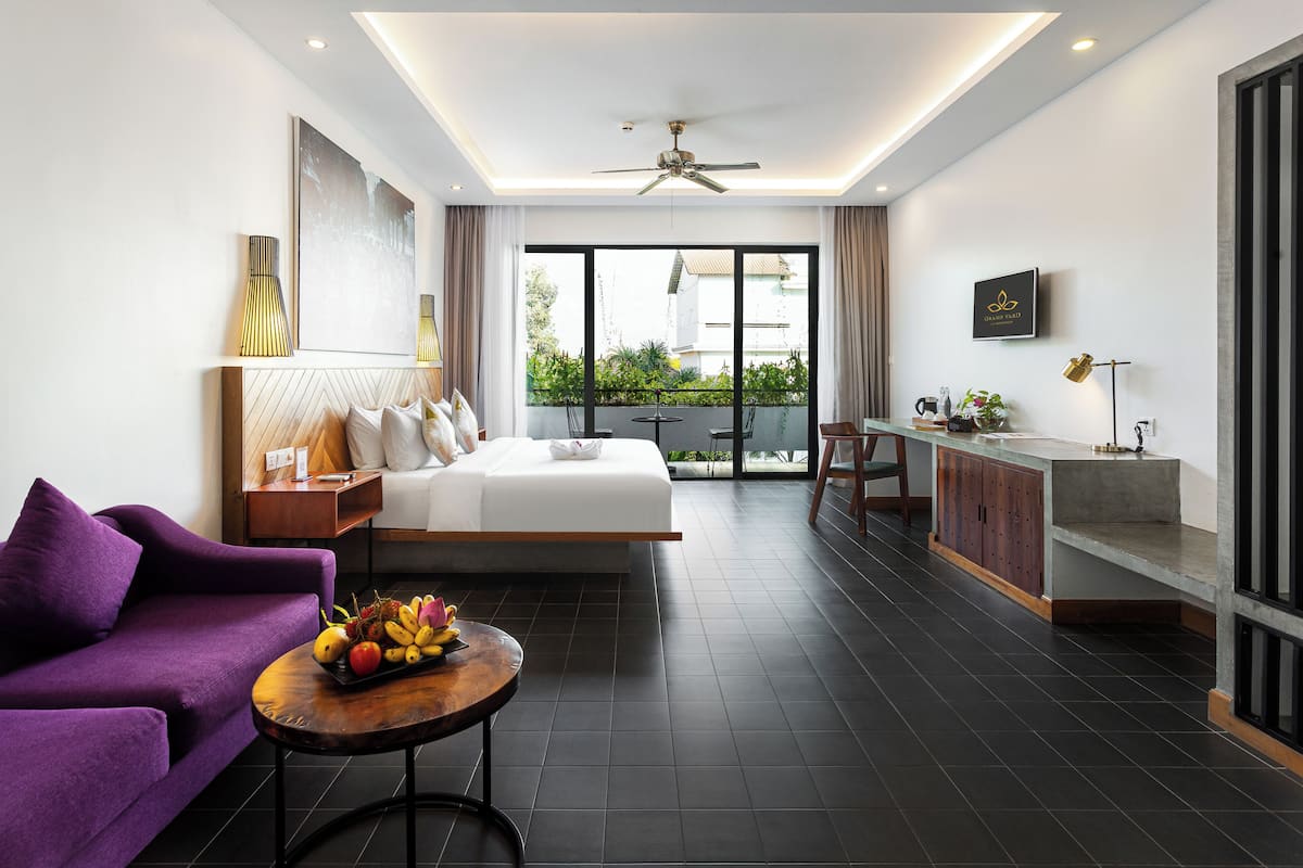 A modern hotel room with a large bed, a purple sofa, a dining area, and a balcony with a view.
