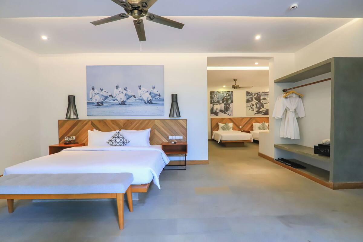 A modern hotel room with a large bed, a ceiling fan, a painting, a wooden headboard, and a walk-in closet with a robe hanging.