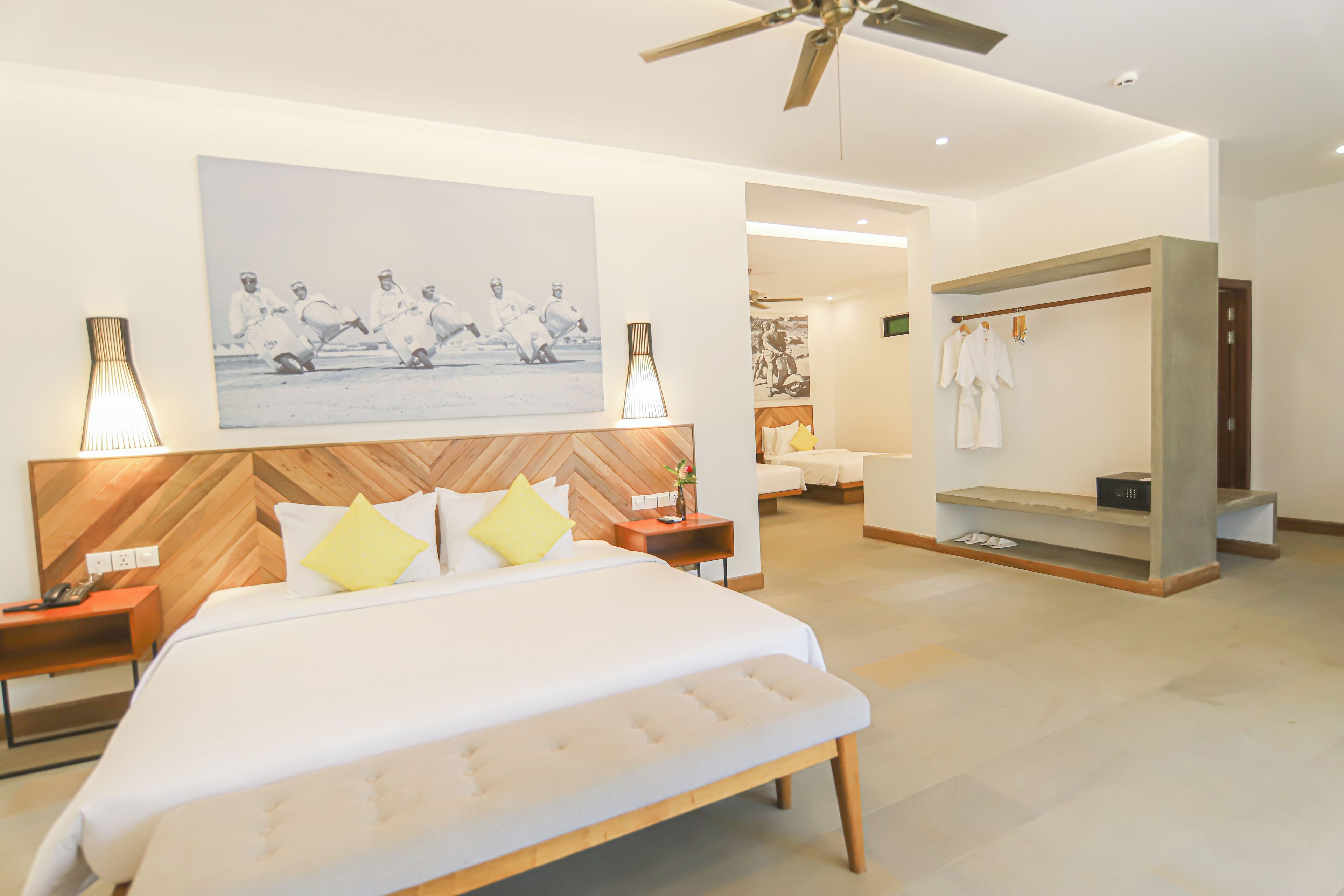 A modern hotel room with a large bed, a wooden headboard, a ceiling fan, and a wardrobe with hanging clothes.