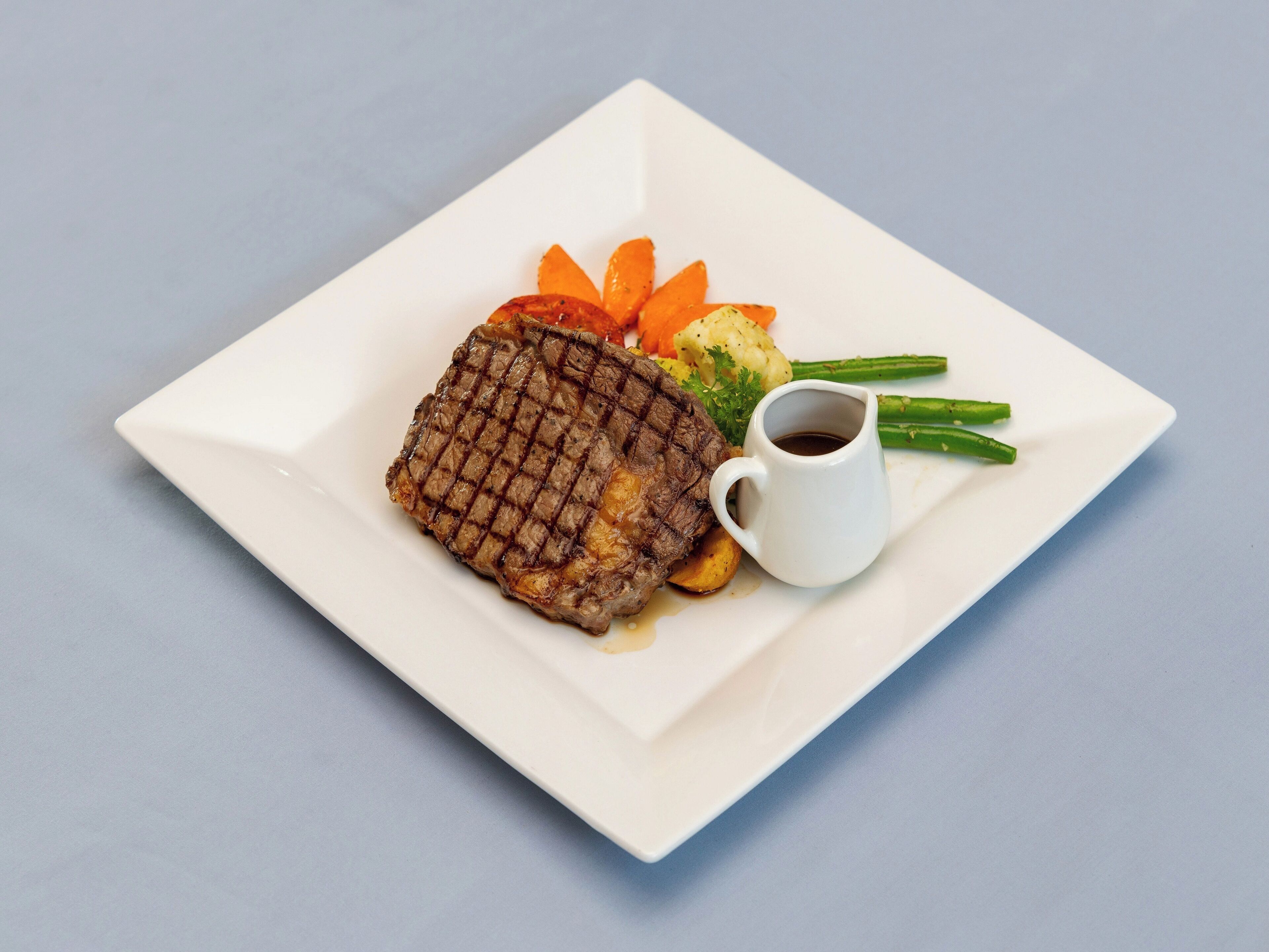 A square white plate with a grilled steak, roasted vegetables, and a small cup of sauce.