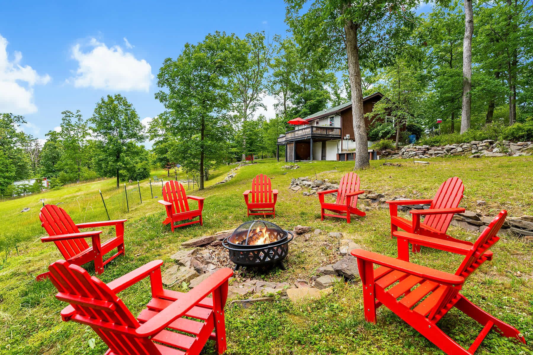 A fire pit with red Adirondack chairs arranged around it, a stone wall, and a house with a balcony in a wooded area.