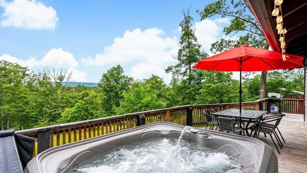 Un bain à remous sur une terrasse en bois, agrémenté d’un parasol rouge, entouré de verdure et sous un ciel dégagé.