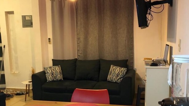 En kompakt opholdsstue med spisebord, røde stole, en sort sofa med mønstrede puder, en rød hængende lampe og et hvidt komfur med kaffemaskine.