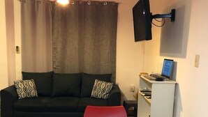 En kompakt opholdsstue med en sofa, et spisebord med røde servietter, et fjernsyn monteret på væggen og et lille skrivebord med en bærbar computer.