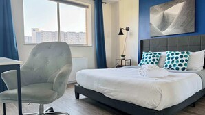 A hotel room with a bed, a chair, a desk, a window with curtains, a lamp, and a wall art piece. - Historic Condo in Atlanta with Skyline View (Atlanta)