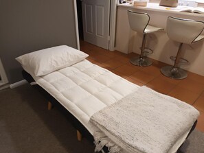 A single bed with a white mattress and pillow, a white throw blanket, and two bar stools in a small room with a tiled floor.