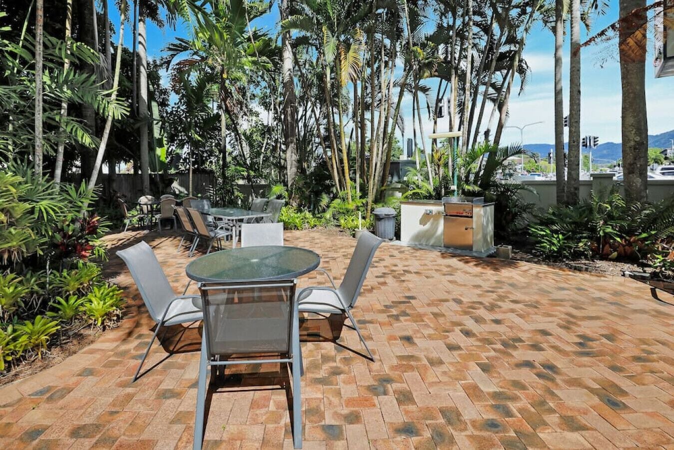An outdoor patio with a round table and chairs, surrounded by lush tropical plants and a brick floor.