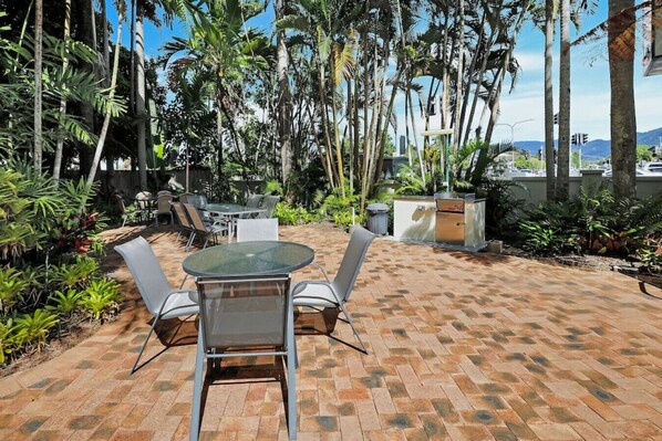 An outdoor patio with a round table and chairs, surrounded by lush tropical plants and a brick floor.