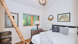A bedroom with a bed, a nightstand, a lamp, a window, a mirror, a wooden ladder, and a wall-mounted wreath.
