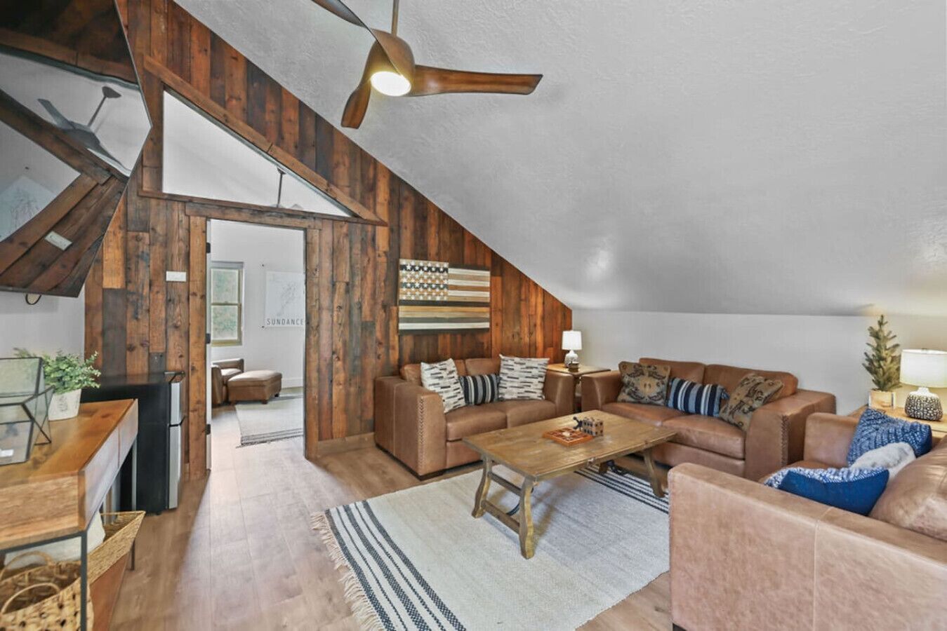 A loft-style living room with exposed wooden beams, a ceiling fan, and a large window.