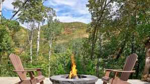 A circular fire pit with two Adirondack chairs on either side, set on a concrete patio with a backdrop of trees and hills.