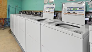 A row of white front-loading washers with control panels, a blue cart, and a green wall with informational posters. - Mathis Motel (Mathis)