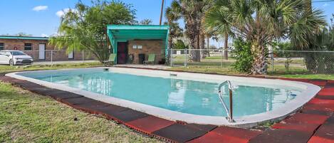 Piscina com deck vermelho e preto, rodeada por palmeiras e um pequeno edifício com telhado verde.