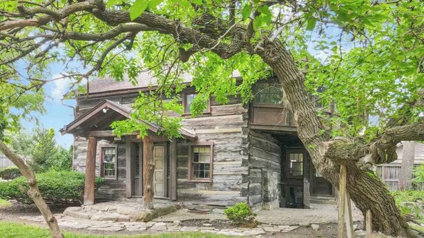 A rustic log cabin with a stone foundation, a front porch, and a large tree in the foreground. - The City Cabin - Unique and Historic! (Columbus)