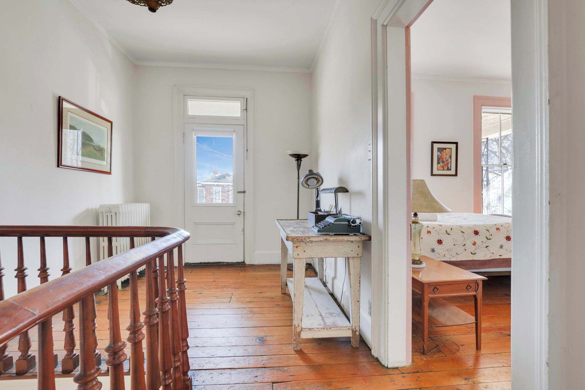 A hallway with a wooden staircase, a white door, a radiator, a table with a lamp, and a bed with floral bedding.
