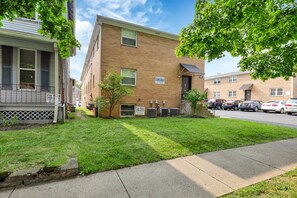 A multi-story brick building with a 'For Rent' sign, a small lawn, and parked cars on the street.