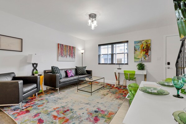 A modern living room with a sofa, coffee table, and a colorful rug. There is a dining area with a table set for two and a window with blinds.