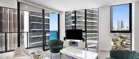 Una sala de estar moderna con sofá, sillas, una mesa de centro y televisor. Amplias ventanas ofrecen vistas a un paisaje urbano y al océano.