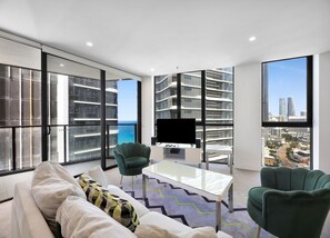 A modern living room with a sofa, chairs, a coffee table, and a TV. Large windows offer views of a cityscape and the ocean. - Wonderful 2-Bed with Pool, Gym & Scenic Views (Surfers Paradise)