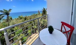 A balcony with a white table and red chair, overlooking a tropical landscape with palm trees and the ocean. - Red Flamboyan Guesthouse (Rincon)