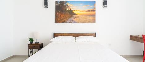 A bedroom with a large bed, a nightstand with a lamp, a desk with a chair, and a wall-mounted picture of a sunset over a beach.